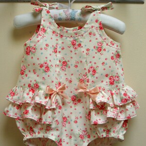 May include: A white baby onesie with a pink floral print and a ruffled bottom. The onesie has two pink bows on the front and is hanging on a white hanger.