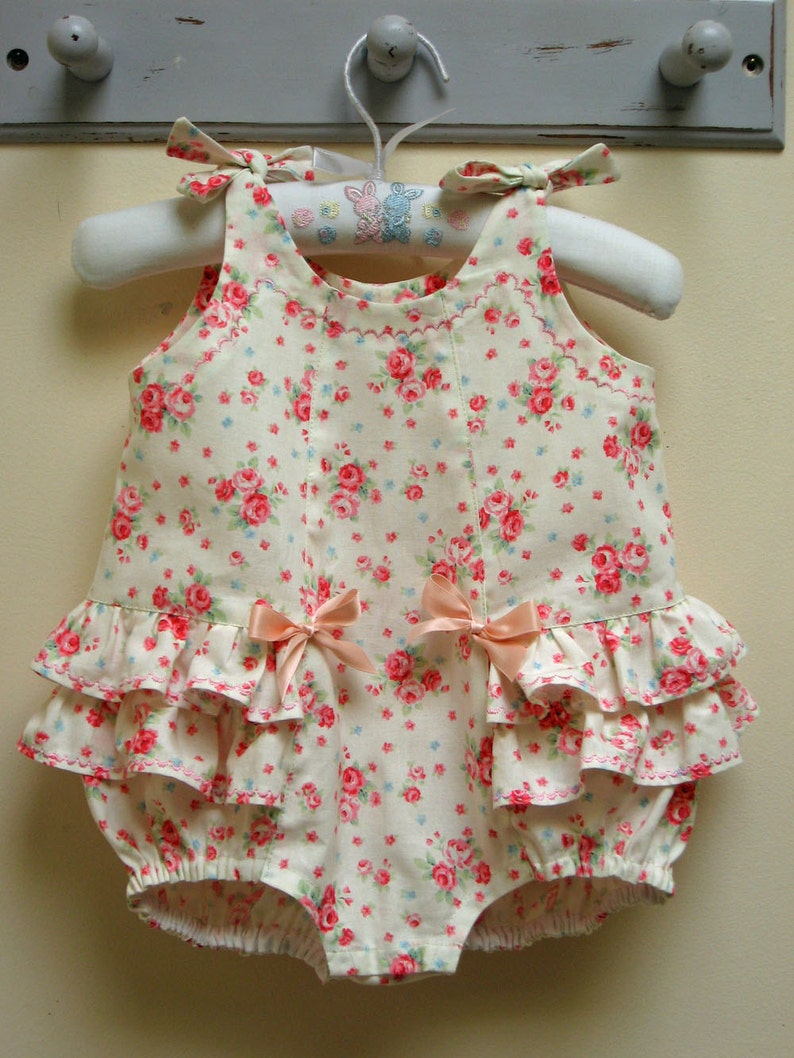 May include: A white baby onesie with a pink floral print and a ruffled bottom. The onesie has two pink bows on the front and is hanging on a white hanger.