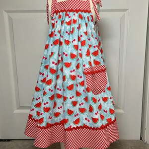 May include: A sleeveless dress with a red and white gingham bodice and a blue and white watermelon print skirt. The dress has a red gingham pocket on the right side and a red gingham trim around the bottom.