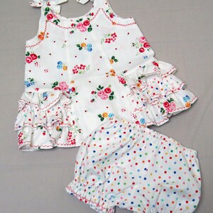 May include: A white sleeveless dress with a floral print and a ruffled hem. The dress is paired with white bloomers with colorful polka dots.