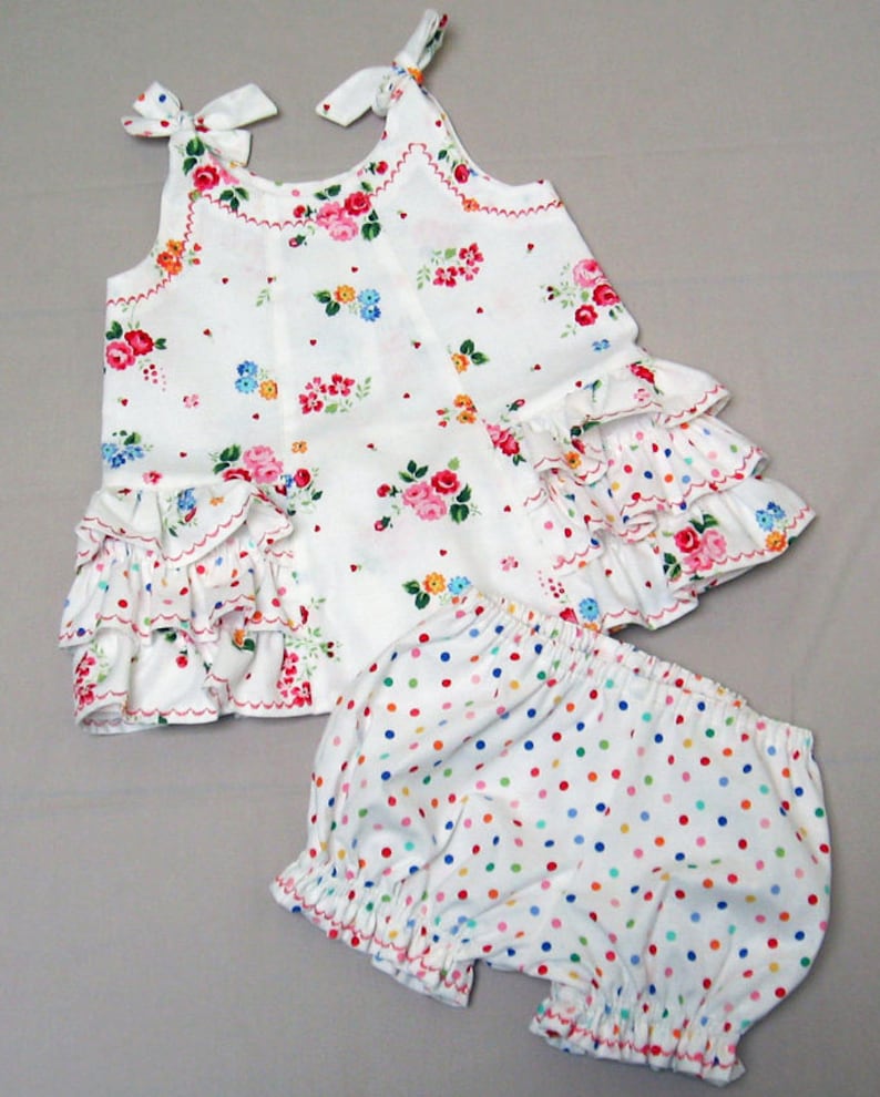 May include: A white sleeveless dress with a floral print and a ruffled hem. The dress is paired with white bloomers with colorful polka dots.