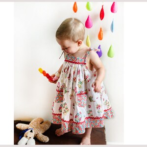 May include: A young girl wearing a white dress with a colorful floral pattern and red trim. She is holding a colorful toy and standing in front of a white wall with a rainbow of felt raindrops hanging from the ceiling.