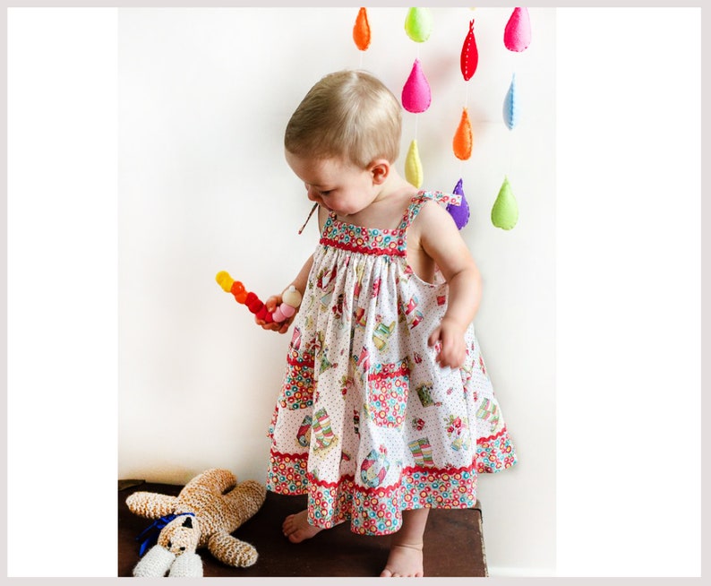 May include: A young girl wearing a white dress with a colorful floral pattern and red trim. She is holding a colorful toy and standing in front of a white wall with a rainbow of felt raindrops hanging from the ceiling.