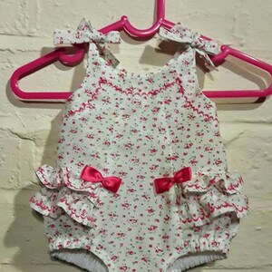 May include: A white baby onesie with a floral print and pink bows. The onesie has a ruffled bottom and is hanging on a pink hanger.