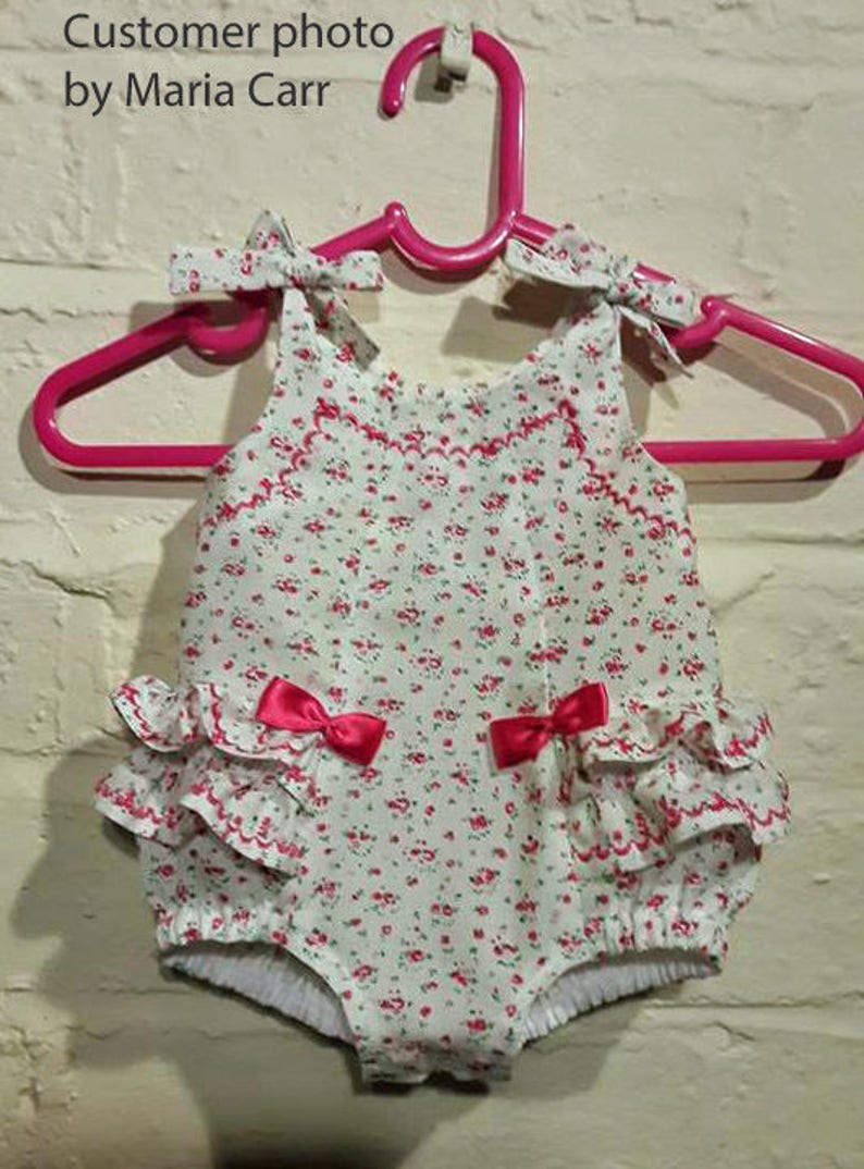 May include: A white baby onesie with a floral print and pink bows. The onesie has a ruffled bottom and is hanging on a pink hanger.
