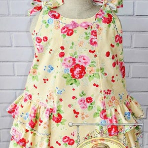 May include: A yellow sleeveless dress with a floral print and a pink bow. The dress has a ruffled hem and is layered with three tiers of ruffles. The dress is on a white mannequin against a white brick wall. The image includes the text "Customer photo by Nicole's Needle".
