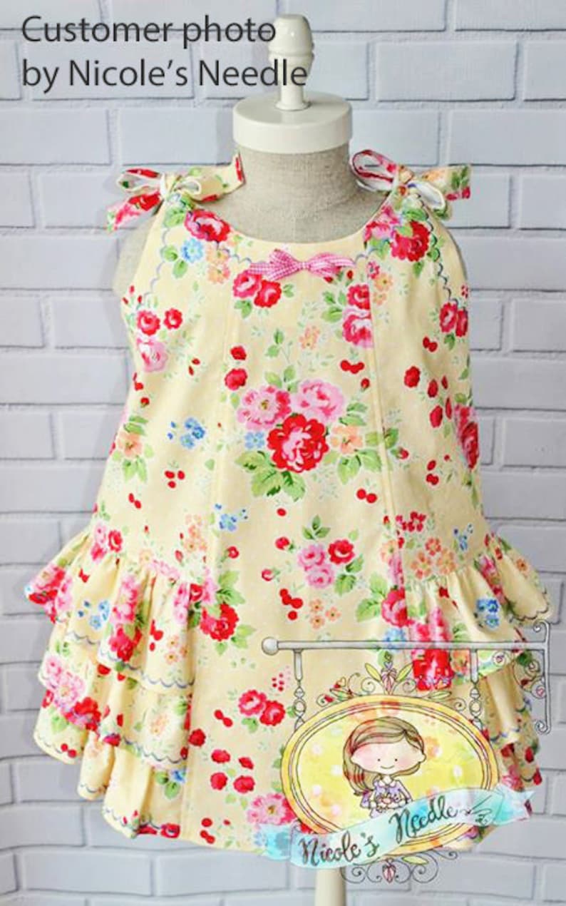 May include: A yellow sleeveless dress with a floral print and a pink bow. The dress has a ruffled hem and is layered with three tiers of ruffles. The dress is on a white mannequin against a white brick wall. The image includes the text "Customer photo by Nicole's Needle".