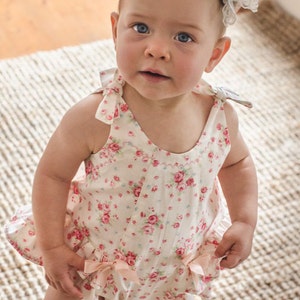 May include: A baby girl wearing a white floral print romper with pink bows. The romper has a ruffled hem and a scoop neckline. The baby is wearing a headband with a white and gray flower.