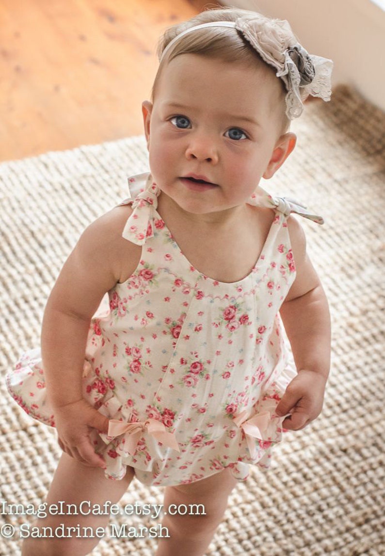 May include: A baby girl wearing a white floral print romper with pink bows. The romper has a ruffled hem and a scoop neckline. The baby is wearing a headband with a white and gray flower.
