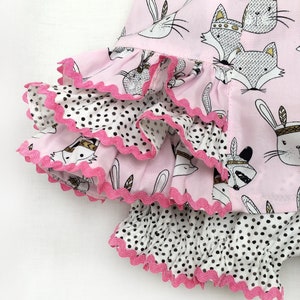 May include: Pink and white fabric with a black and white polka dot pattern. The fabric has a ruffled edge and features a print of animals wearing headdresses.