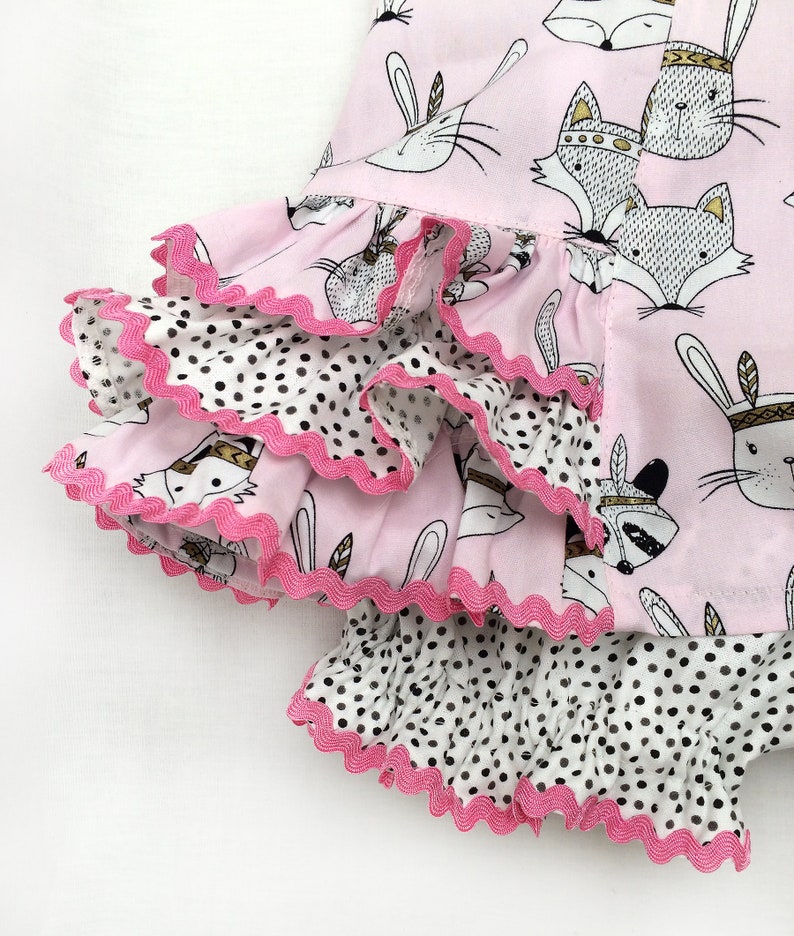 May include: Pink and white fabric with a black and white polka dot pattern. The fabric has a ruffled edge and features a print of animals wearing headdresses.