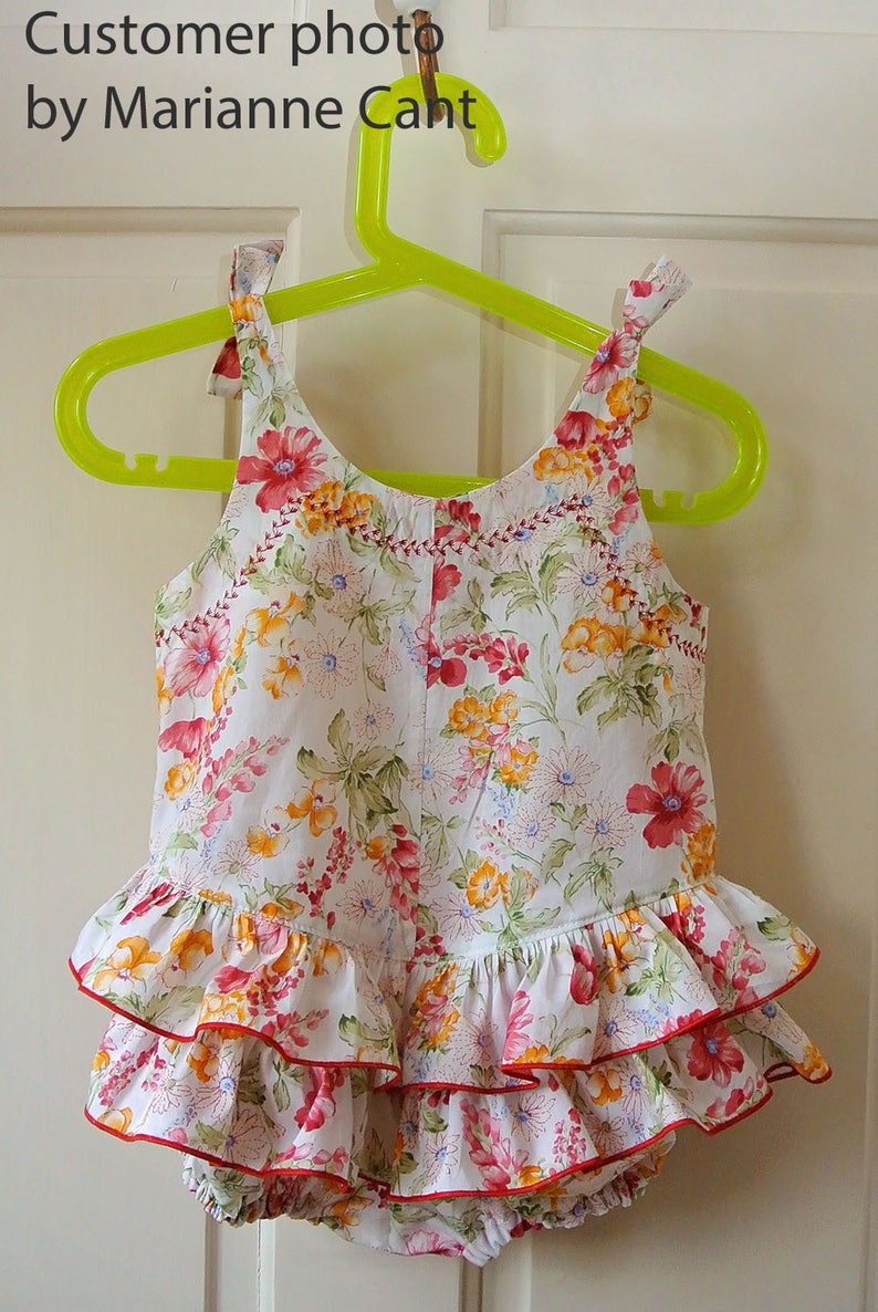 May include: A white baby onesie with a floral print in shades of pink, yellow, and green. The onesie has a ruffled skirt with three layers and a red trim.
