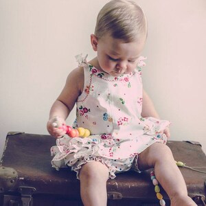 May include: A young child wearing a white dress with floral print sits on a vintage suitcase. The child is playing with a colorful wooden toy.