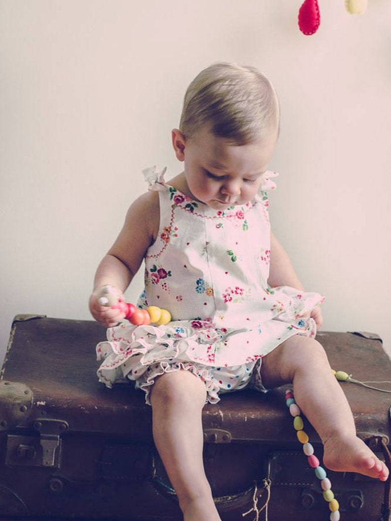 May include: A young child wearing a white dress with floral print sits on a vintage suitcase. The child is playing with a colorful wooden toy.