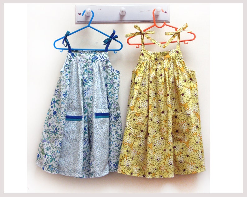 May include: Two sleeveless dresses hanging on hangers. The dress on the left is white with a blue floral pattern. The dress on the right is yellow with a black floral pattern.