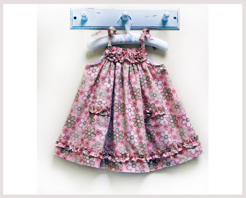 May include: A pink and green floral print dress with ruffles around the neckline and hem. The dress has two pockets on the front.