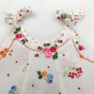 May include: White sleeveless dress with a floral print and pink heart accents. The dress has a scalloped neckline and pink ties at the shoulders.