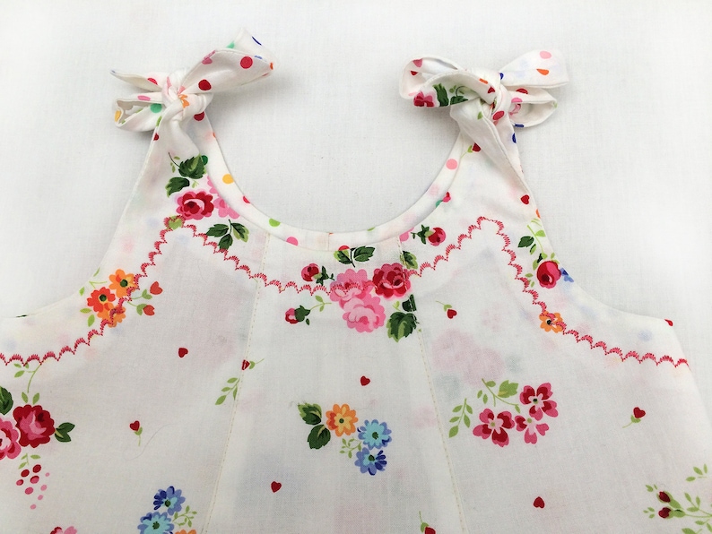 May include: White sleeveless dress with a floral print and pink heart accents. The dress has a scalloped neckline and pink ties at the shoulders.