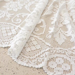 May include: A white lace fabric featuring an intricate floral pattern with a grid-like mesh background. The textile is folded over, revealing the detailed needlework and scalloped edges of the delicate, semi-sheer material with its repeating motifs.