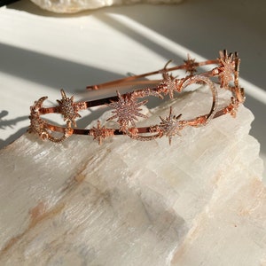 Könnte beinhalten: Ein roségoldenes Stirnband mit einem himmlischen Design aus Sternen und Mondsicheln. Das Stirnband ist mit funkelnden Strasssteinen verziert.