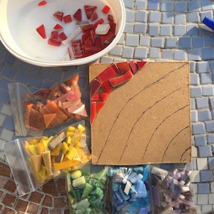 May include: A close-up of a mosaic art project in progress. A white bowl filled with red and white glass tiles sits next to a piece of plywood with a rainbow design drawn on it. Four plastic bags filled with different coloured glass tiles are arranged below the plywood.