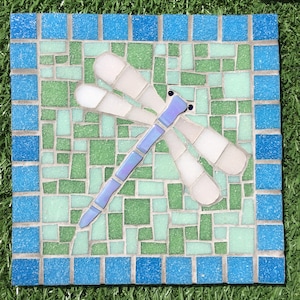 May include: Square mosaic tile with a dragonfly design. The dragonfly has a light blue body and white wings, set against a background of green and blue tiles. The border is a darker blue, creating a decorative garden accent.