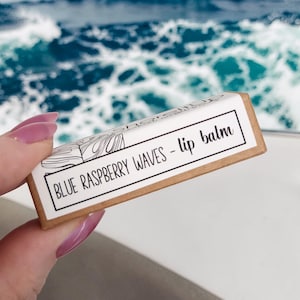 May include: A rectangular lip balm box with the text "BLUE RASPBERRY WAVES - lip balm" in a black font. The box is held in front of a blue ocean background. The box has a white background with a brown border.