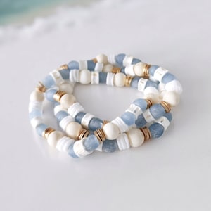 May include: A set of three bracelets with blue and white glass beads and gold spacers. The bracelets are layered on a white surface.