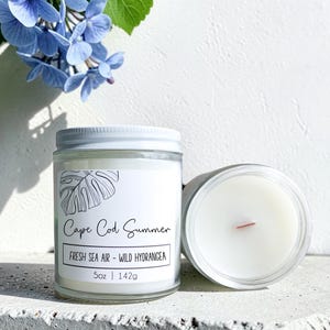 May include: Two clear glass jar candles with white wax and a single wooden wick. The label features a line drawing of a leaf, the text Cape Cod Summer, FRESH SEA AIR - WILD HYDRANGEA, and the weight 5oz | 142g. Blue hydrangea flowers are visible.