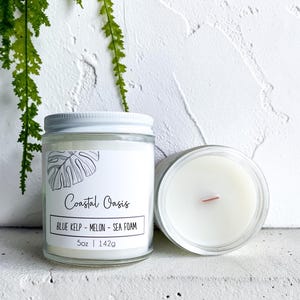 May include: Two clear glass jars containing white wax candles with wooden wicks. The front label features a line drawing of a leaf and the text Coastal Oasis, Blue Kelp - Melon - Sea Foam, and 5oz | 142g. One jar is upright, the other is viewed from above.