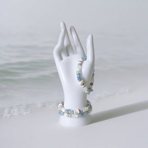 May include: Two beaded bracelets with a beach theme. The bracelets feature a mix of white, tan, and blue beads, including small shells. One bracelet is displayed on a white ceramic hand, with the other bracelet resting below.