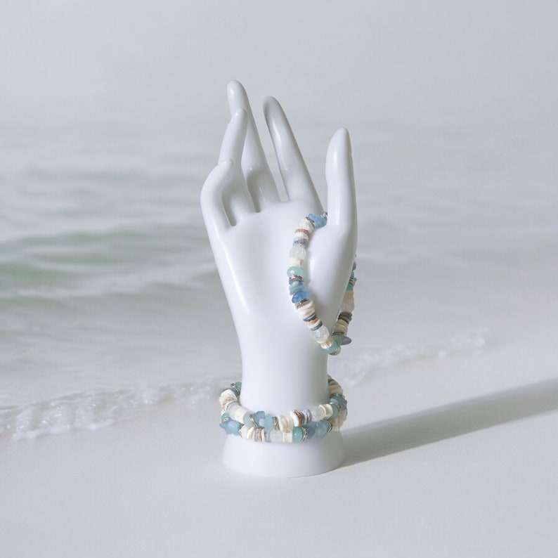 May include: Two beaded bracelets with a beach theme. The bracelets feature a mix of white, tan, and blue beads, including small shells. One bracelet is displayed on a white ceramic hand, with the other bracelet resting below.