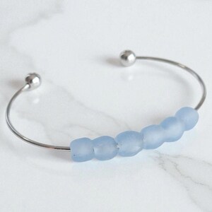 May include: A silver-toned cuff bracelet with a row of light blue beads. The bracelet has a simple, open design with a small silver ball at each end. The beads are a soft, matte finish.