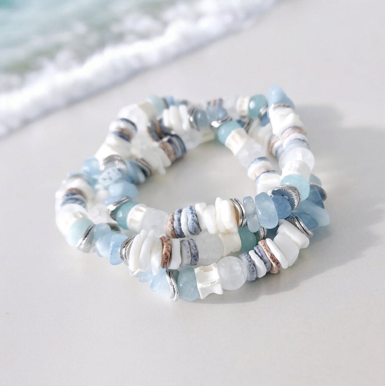 May include: Three stacked bracelets featuring a mix of white, light blue, and brown shell beads. The bracelets have silver-colored metal accents and are arranged on a white surface, with a blurred ocean background.