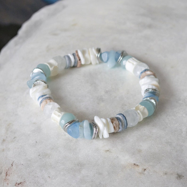 May include: A beaded bracelet featuring a mix of white shell chips, light blue stones, and silver-toned metal accents. The bracelet has a circular shape and is designed to be worn around the wrist. The overall aesthetic is beachy and casual.