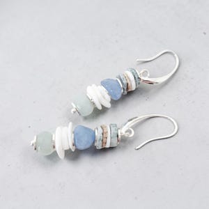 May include: A pair of dangle earrings featuring a combination of white, light blue, and tan beads. The earrings have a silver-colored hook closure. The beads are arranged in a vertical pattern, creating a unique and stylish accessory.