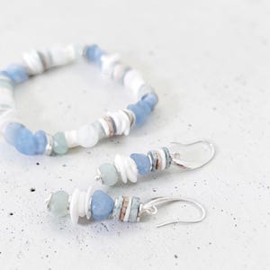 May include: A bracelet and matching earrings set. The jewelry features a combination of white, light blue, and silver beads. The earrings have a hook closure. The bracelet and earrings are made with a variety of shapes and sizes of beads.