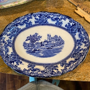 May include: A large, oval, blue and white porcelain serving platter with a raised rim and a floral pattern. The center of the platter features a blue and white illustration of a castle and surrounding landscape.
