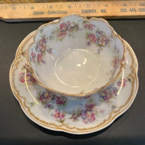 May include: A white porcelain teacup and saucer set with a floral pattern in shades of pink, yellow, and blue. The set has a gold trim around the edges.