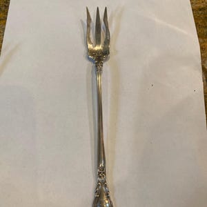 May include: A silver fork with an ornate handle and three tines. The fork is on a white background.