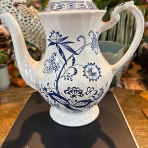 May include: White ceramic teapot with a blue floral design. The teapot has a curved spout, a handle, and a lid with a finial. The floral pattern includes stylized flowers and leaves. The teapot is sitting on a black surface.