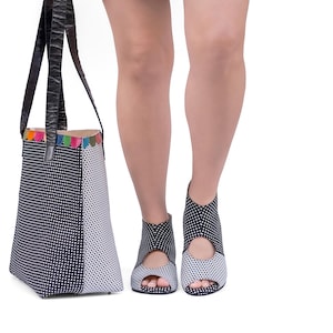 Op de afbeelding: Een zwart-witte stippen tote bag met kleurrijke afwerking en zwarte leren handvatten. De tas wordt gedragen door iemand die zwart-witte stippen open-toe schoenen draagt.