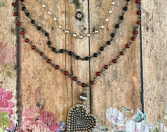 Puede incluir: Un collar de varias cadenas en tono bronce antiguo adornado con cuentas transparentes, crema, negras y rojo oscuro. El diseño incluye un colgante central en forma de corazón con una superficie texturizada y un pequeño dije circular.