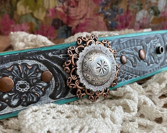 May include: A turquoise and black leather bracelet with a silver and copper floral design. The bracelet has a decorative silver and copper centerpiece with a starburst pattern.