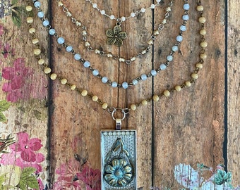 Puede incluir: Un collar de varias capas con una cadena plateada, cuentas azules y beige, y un colgante rectangular con un diseño floral.