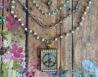 Puede incluir: Un collar de varias capas con un colgante de signo de paz. El collar presenta múltiples hilos de cuentas en crema, azul claro y morado. El colgante de signo de paz es dorado con un diseño blanco y negro.