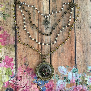 Könnte beinhalten: Eine mehrlagige Halskette mit einem großen, runden Anhänger. Der Anhänger ist aus Metall und hat ein florales Design mit grünen, rosa und weißen Akzenten. Die Halskette besteht aus braunen, weißen und goldenen Perlen und Ketten.