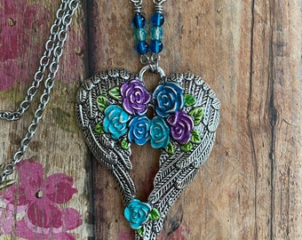 Könnte beinhalten: Eine silberfarbene Metall-Halskette mit einem herzförmigen Anhänger, der aus zwei Flügeln besteht. Die Mitte ist mit emaillierten violetten, blauen und türkisfarbenen Rosen sowie grünen Blättern verziert. Die Kette enthält kleine blaue und türkise Glasperlen.
