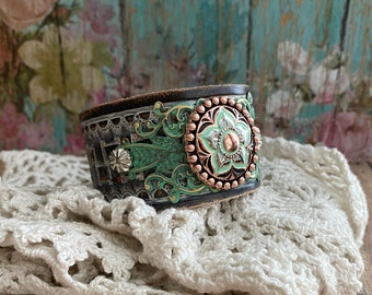 May include: A wide leather cuff bracelet with a distressed black finish. It features intricate green and copper-colored floral designs and a central decorative element with a gemstone. The cuff is resting on a cream-colored crocheted cloth.