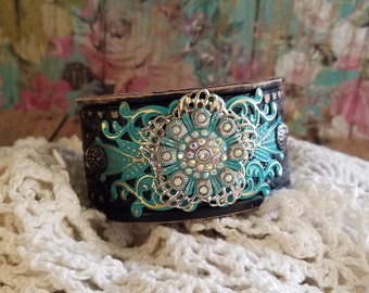 May include: A black leather cuff bracelet with a turquoise and gold filigree design and a large rhinestone embellishment.